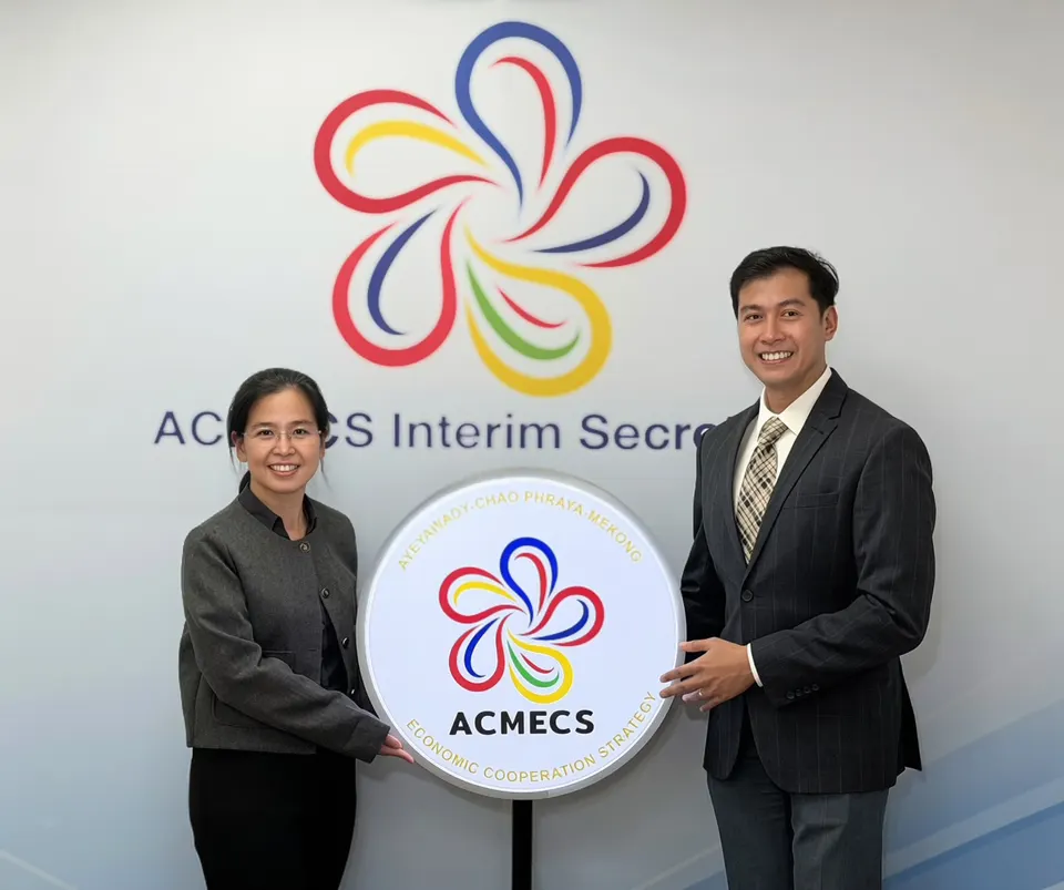 A woman and a man in formal wear hold an illuminated round ACMECS logo sign in front of a wall bearing the ACMECS Interim Secretariat logo
